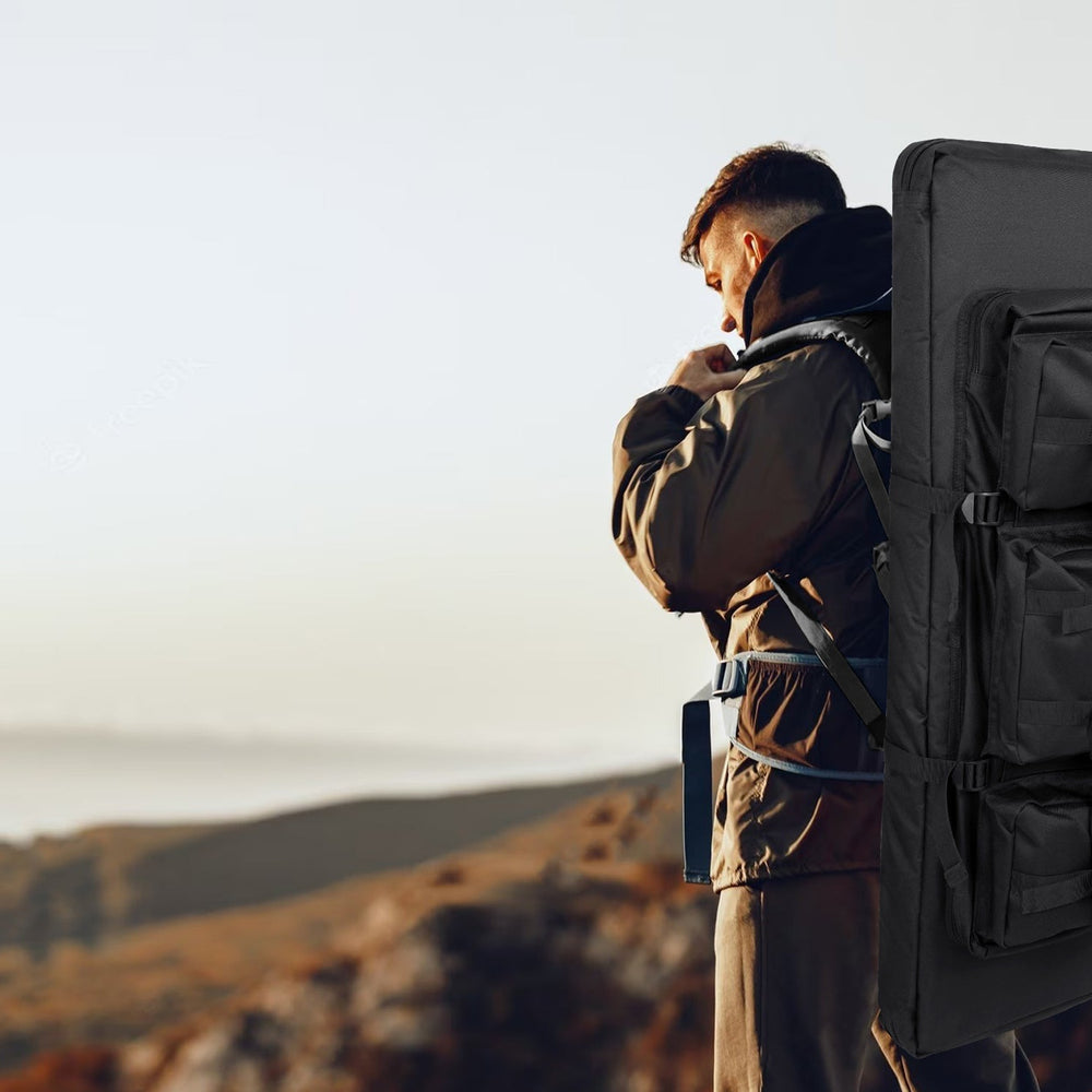 Man wearing a large black tactical rifle case backpack stands on a cliff edge overlooking a hazy landscape
