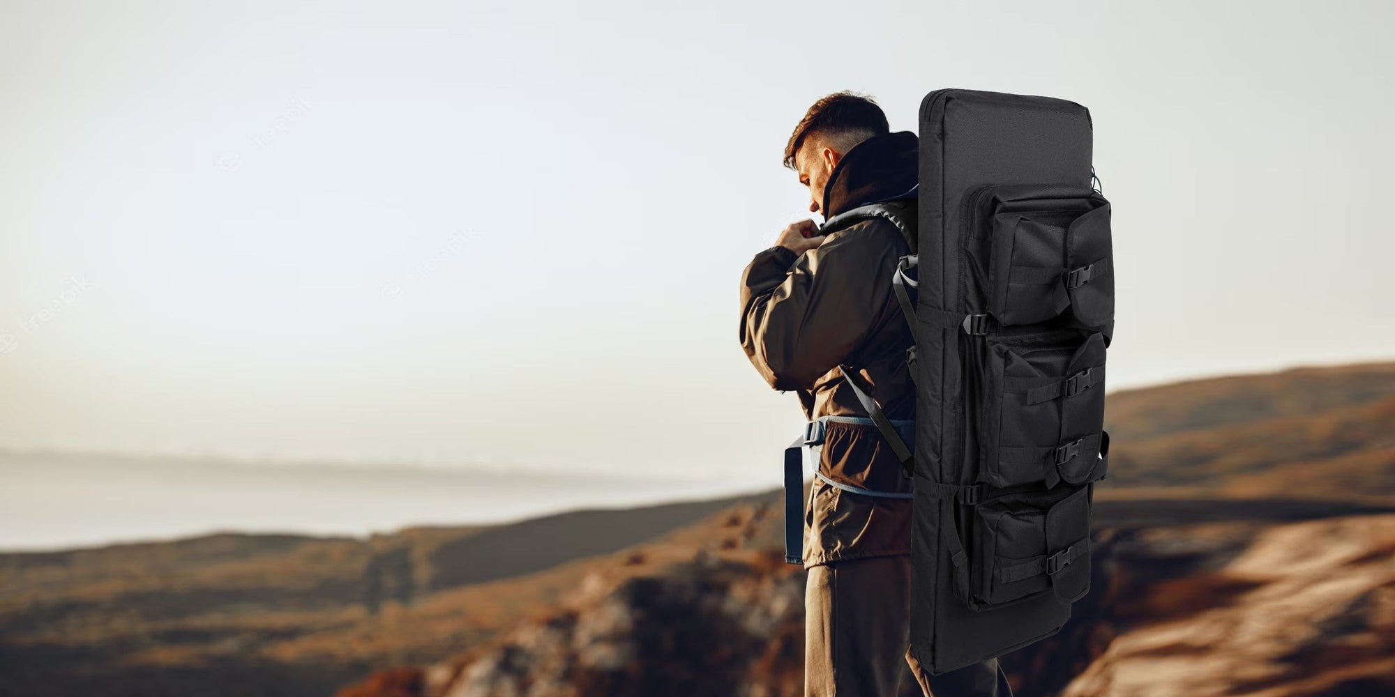 Man wearing a large black tactical rifle case backpack stands on a cliff edge overlooking a hazy landscape